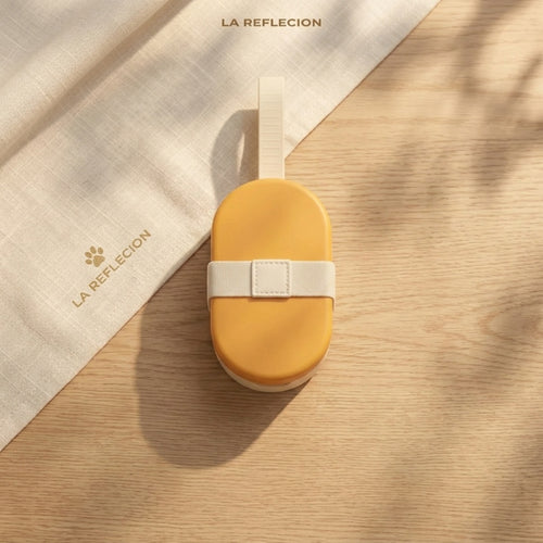 A yellow oval-shaped pet food box with a white strap, placed on a light wood surface next to a beige linen cloth printed with the brand name LA REFLECION.